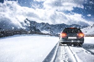Black car driving on snow-covered roads near mountains needing vehicle winterization services.