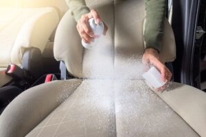 Person spraying a cleaning chemical aerosol can on the tan fabric upholstery of their vehicle in Springfield, IL.