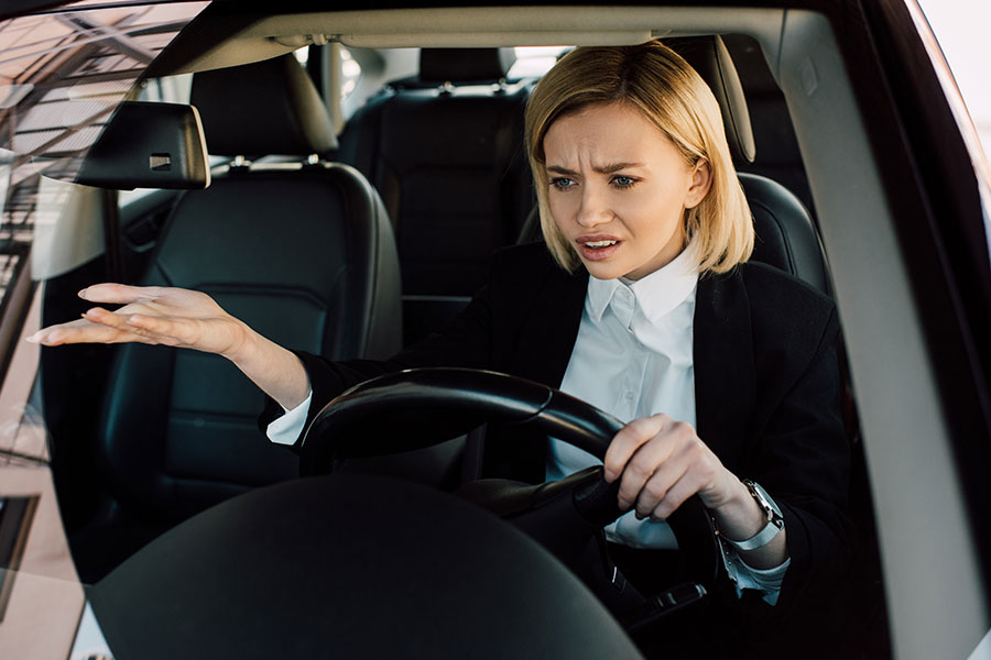 A young blonde woman sits behind the steering wheel of a car in Springfield, IL, gesturing with her hand to signify her frustration and confusion about the loud noises her car makes when she taps the accelerator.