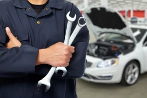 A male mechanic in uniform with his arms crossed holding tools with a car in the background at a shop in Springfield, IL.